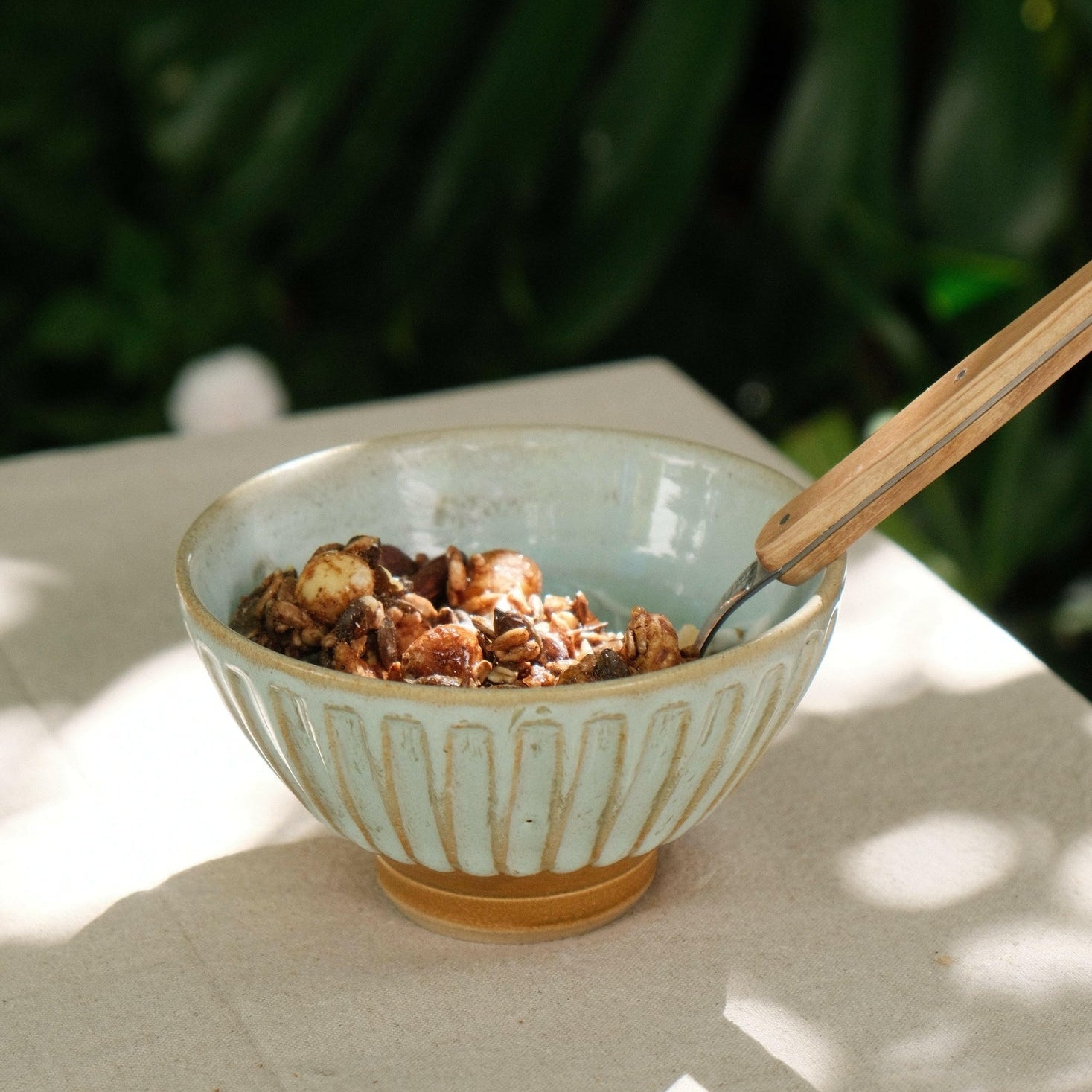 MANTRA LIVING fluted ceramic bowl holding granola with a wooden spoon, on an outdoor table in dappled light. #Color_Desert Oasis