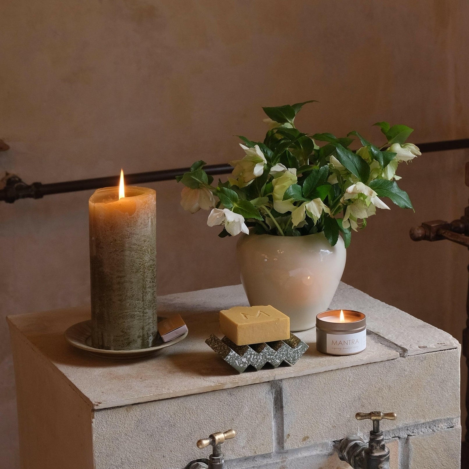 MANTRA LIVING olive green frosted pillar candle lit on a ceramic saucer beside a jug of white hellebores. #Color_Olive
