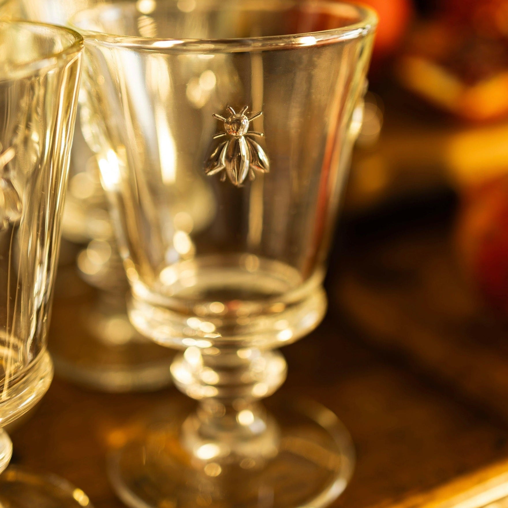 MANTRA LIVING La Rochère bee glass on a brass tray in candlelight, embossed bee motif on a footed clear goblet.