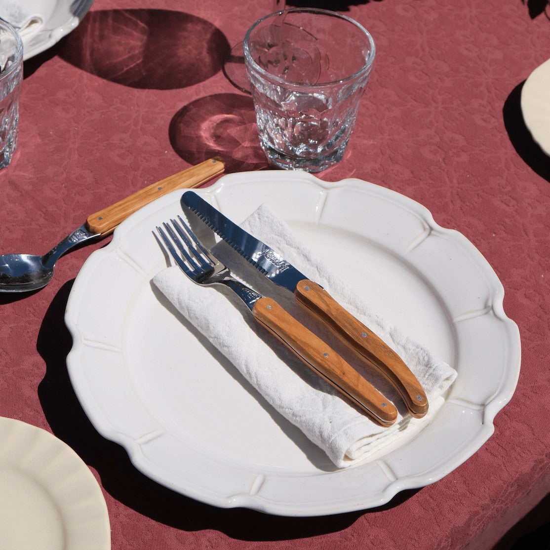 Mantra Living scalloped plate laid with a linen napkin and olive wood handled cutlery on a deep red tablecloth.