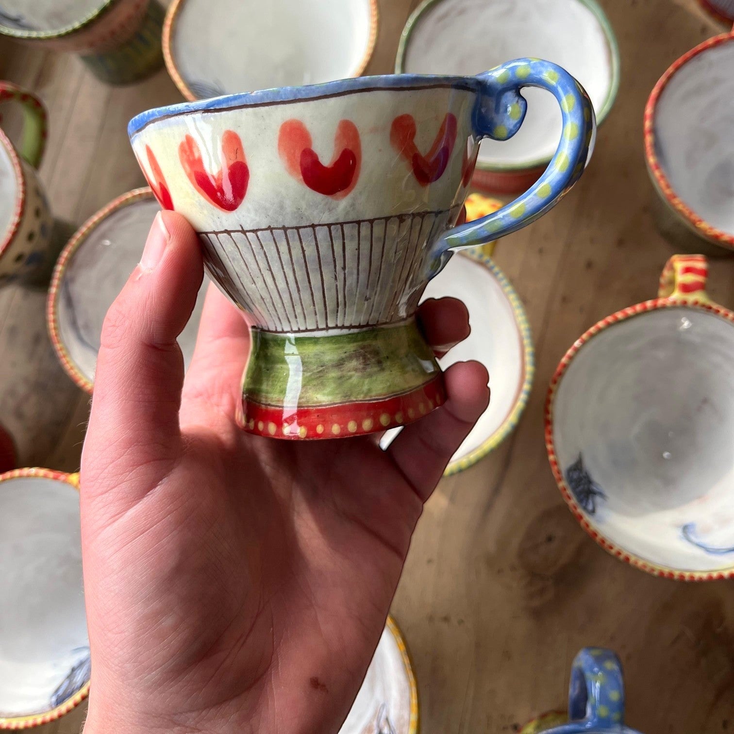 MANTRA LIVING Isabella tea cup with hand-painted red hearts and blue handle, held in hand above other ceramic cups.