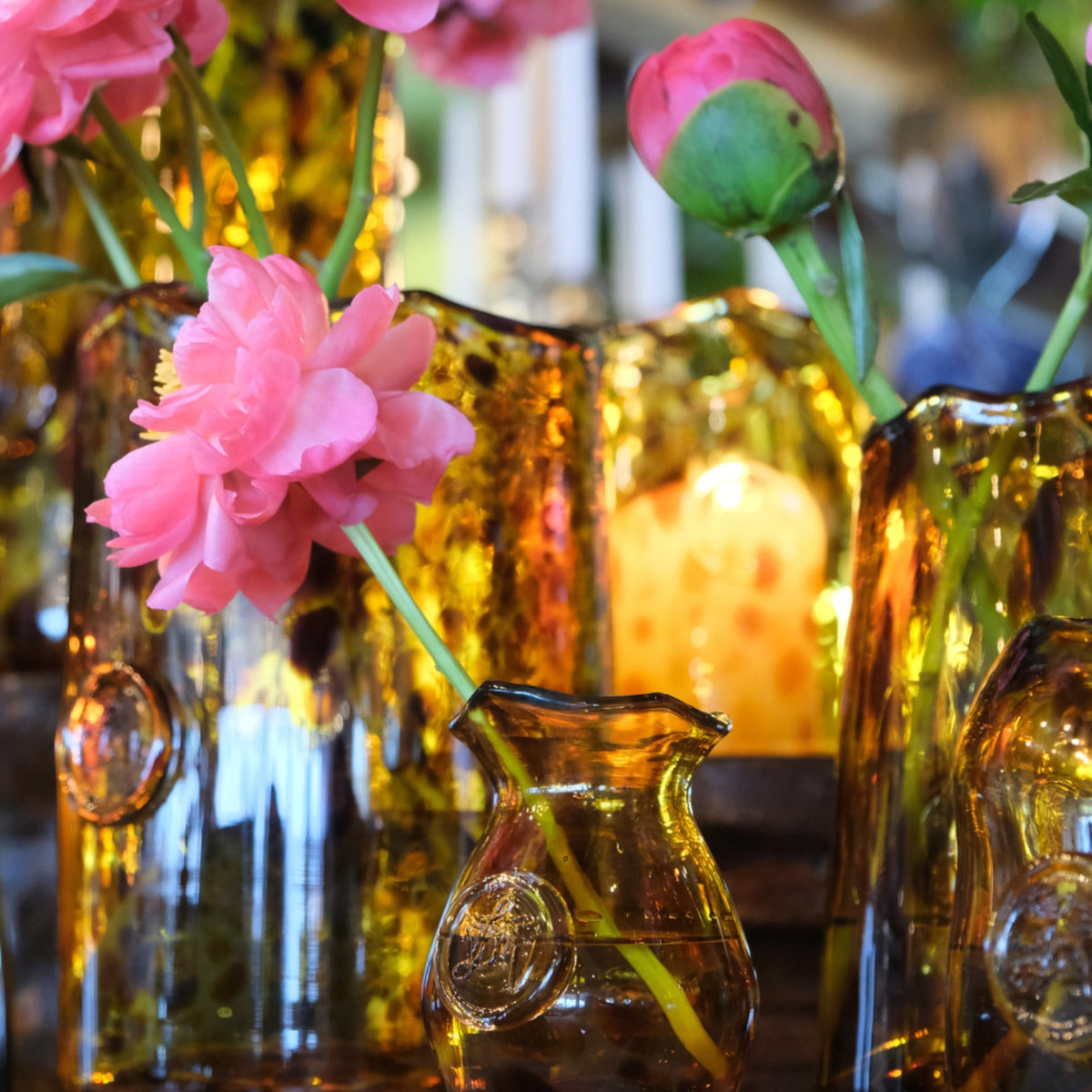 MANTRA LIVING Mia Vase in amber glass with a fluted rim, holding a pink peony stem among other handcrafted glass vases.