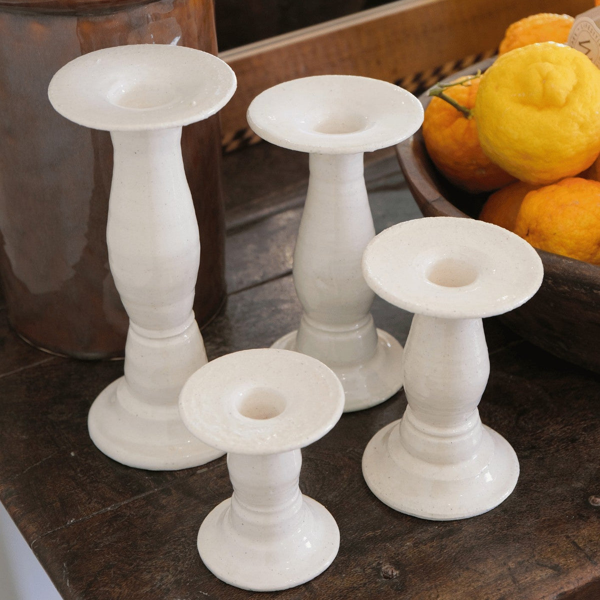 Four Siena candlestick holders in cream Panna ceramic, one of each height, on a wooden table beside a bowl of lemons. #Color_Panna