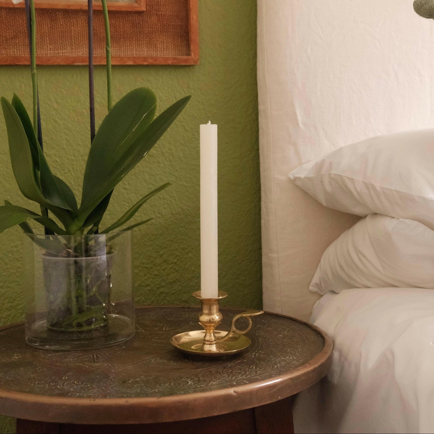 Mantra Living Frosted Dinner Candle in cream, lit in a brass holder on a bedside table beside an orchid. #Color_Cream