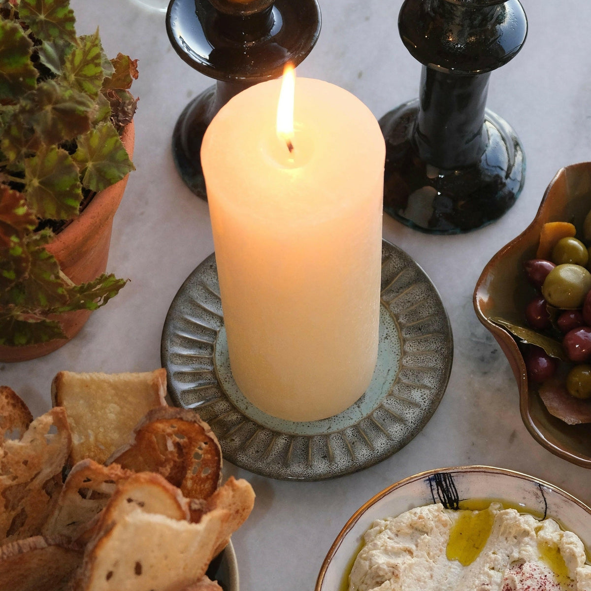 MANTRA LIVING cream frosted pillar candle lit on a fluted saucer on a marble table set with olives, dips and crisps. #Color_Cream