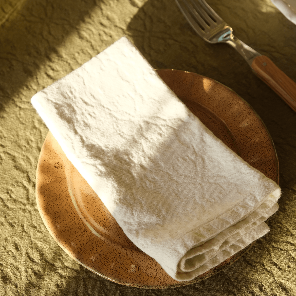 Mantra Living Hibiscus cotton napkin folded on a speckled terracotta plate over an olive linen tablecloth in afternoon light.