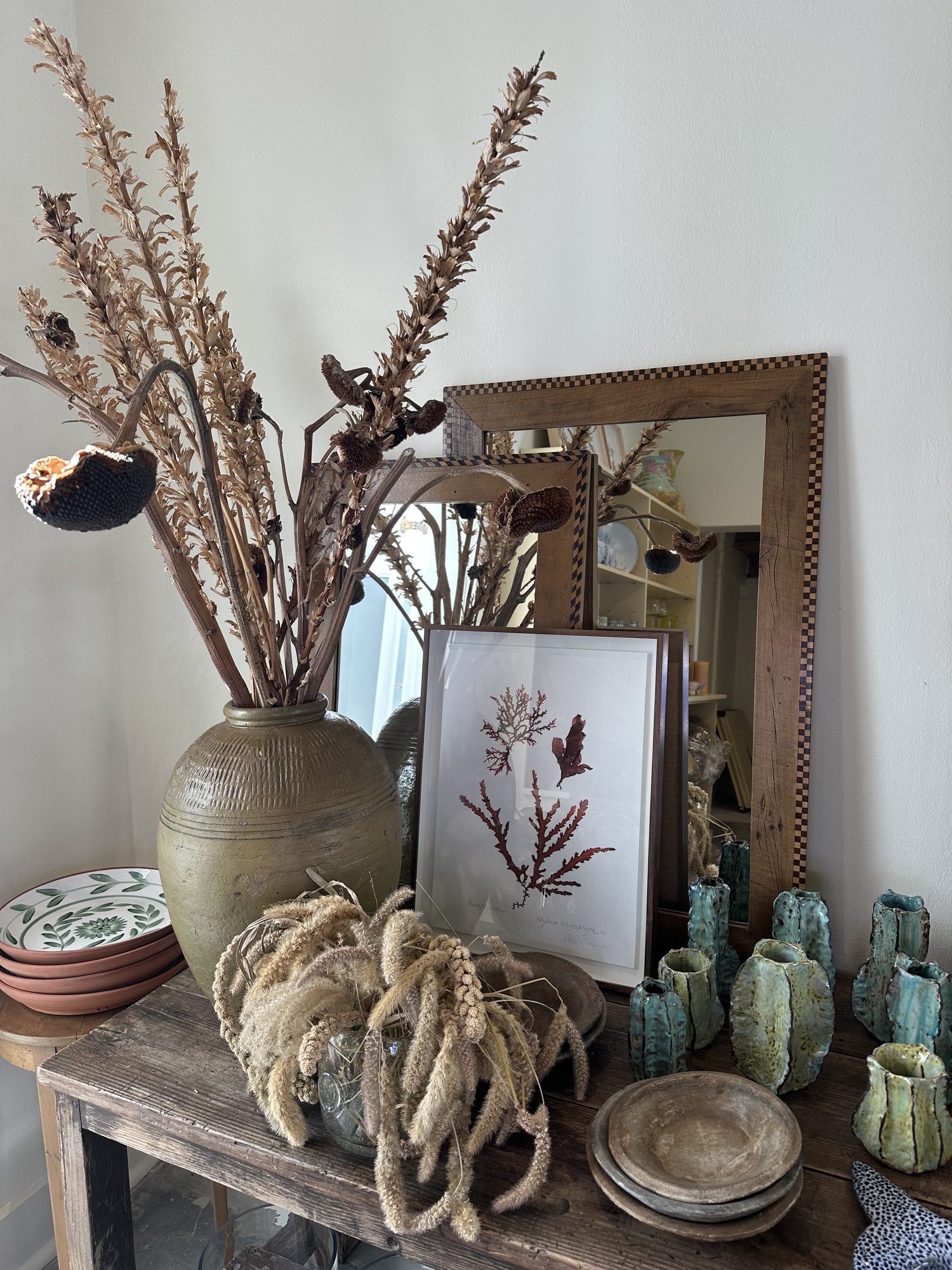 A console styled with dried banksia in a stoneware urn, a framed seaweed print and green ceramics
