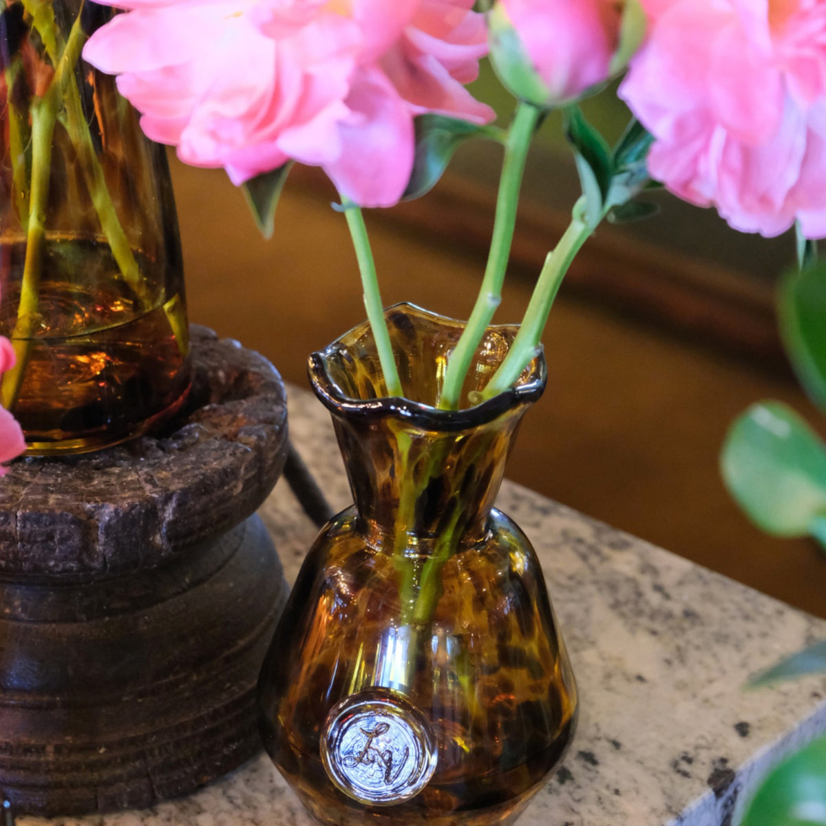 MANTRA LIVING Casa LV Peony Vase in amber glass with ruffled rim and embossed logo, holding pink peony flowers.