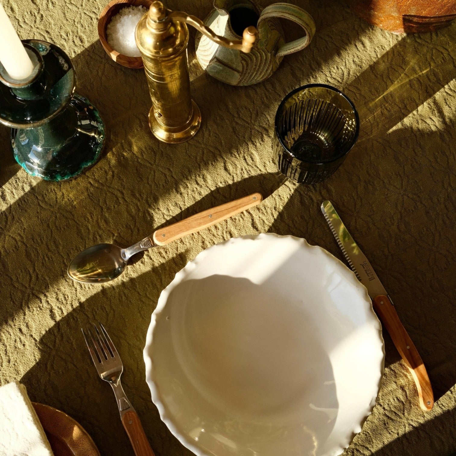 Mantra Living Laguiole olive wood cutlery on an olive linen table with a scalloped white bowl and green glassware.