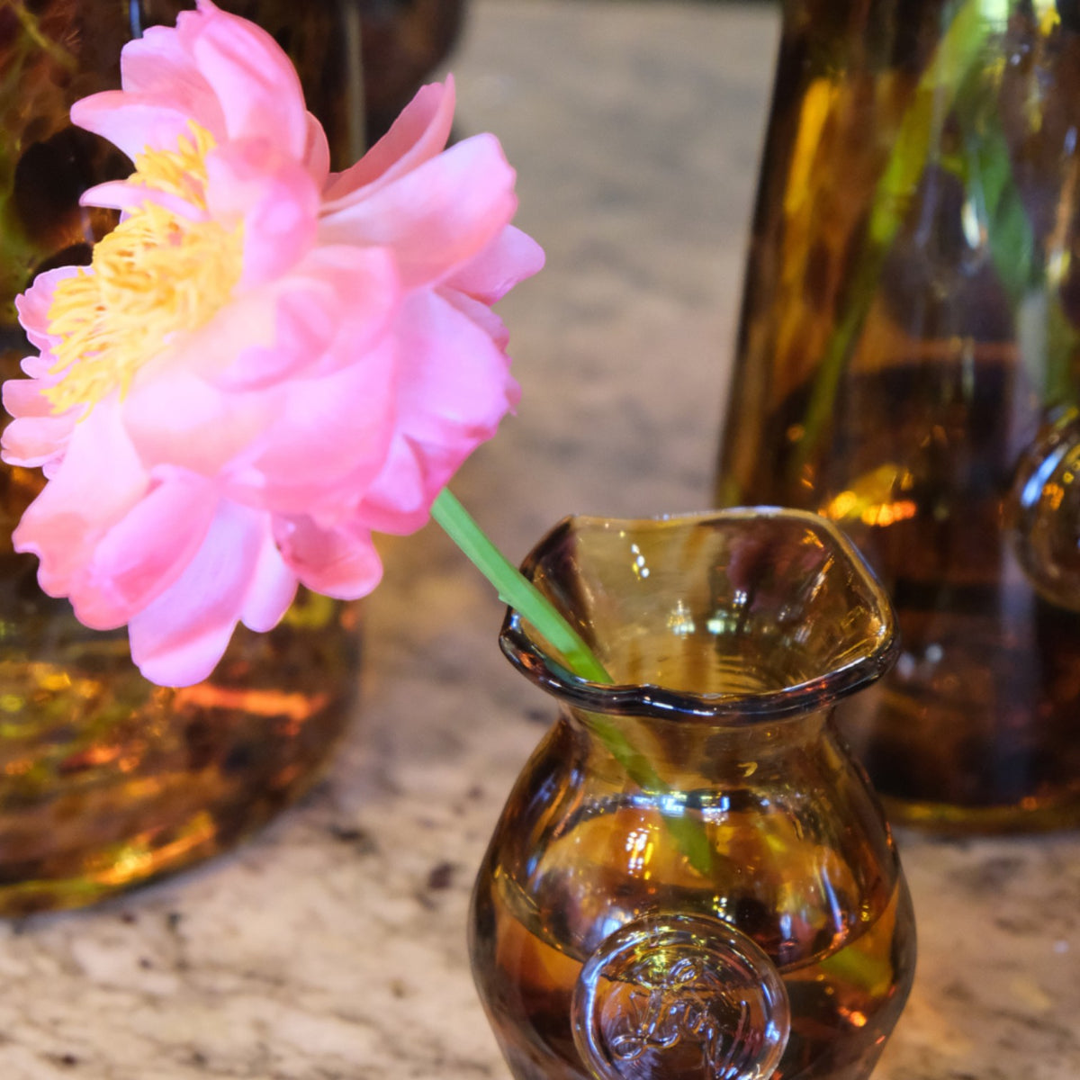 MANTRA LIVING Mia Vase in amber glass with a fluted rim, holding a single pink flower on a marble surface.